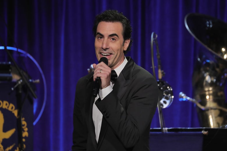 Sacha Baron Cohen holds a microphone on stage at the Friars Club in New York City.